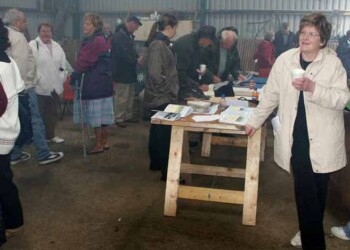 Someone has to hold the table up Group of people in a barn talking and drinking tea with lady leaning on table in foreground