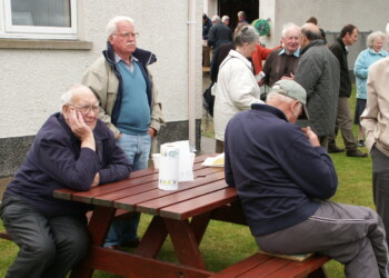 3 men sitting at picnic bench in foreground with larger mixed group behind them